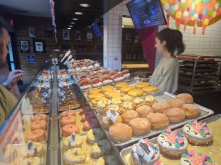 A person standing behind a counter with trays of doughnuts

Description automatically generated