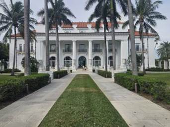 A white building with columns and a walkway with palm trees with Flagler Museum in the background

Description automatically generated